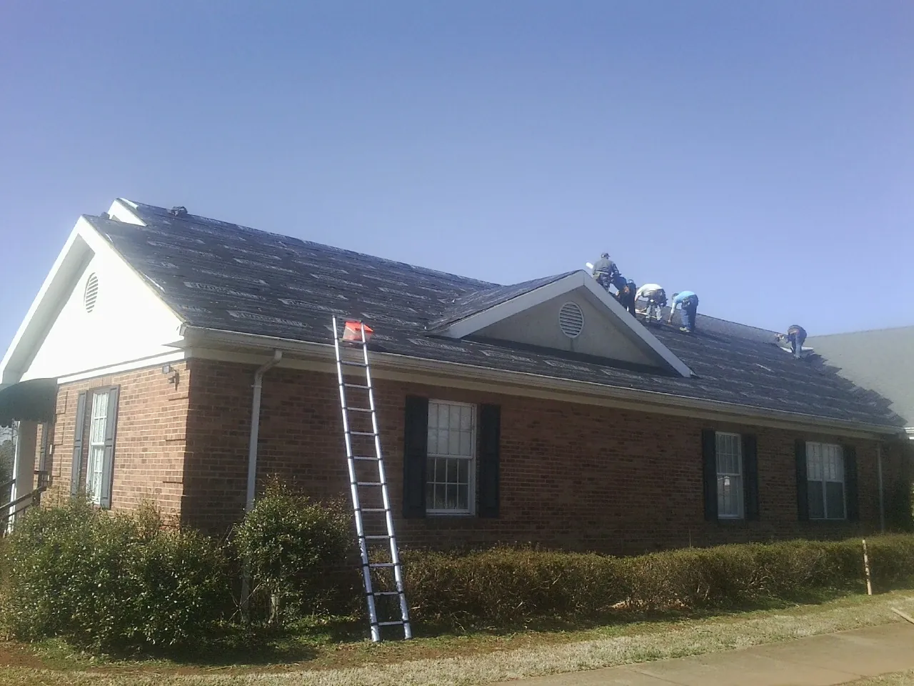 Crew tearing shingles off a brick ranch roof with a ladder set — Emergency Roof Repair work by our crew in Ligonier