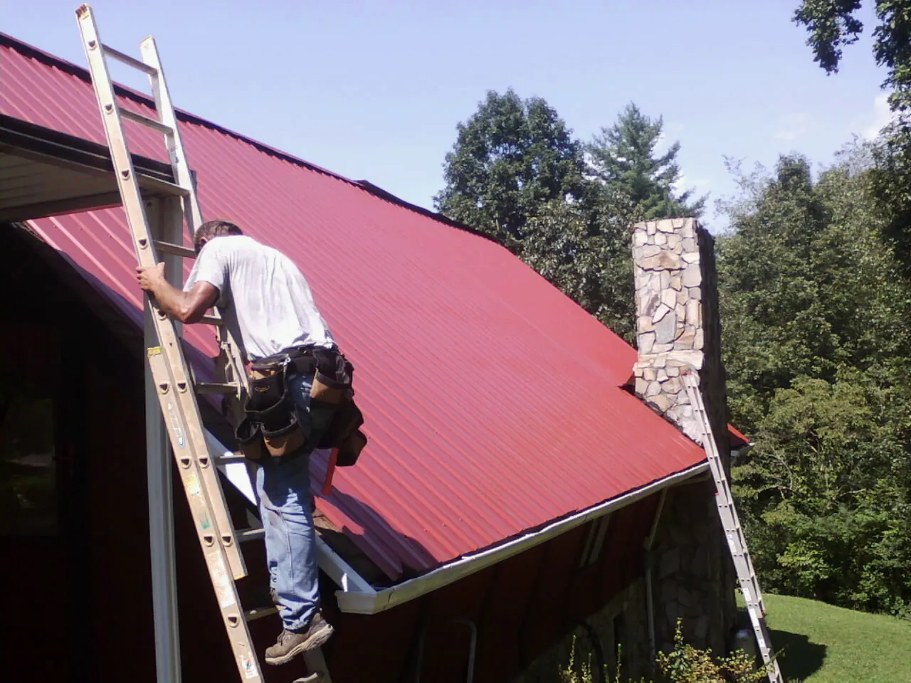 Roofer on a ladder beside a red metal roof and stone chimney — Soffit & Fascia Repair by Roofing Brothers in Ligonier
